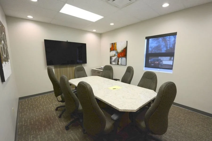 Conference room with oval marble table, eight chairs, wall-mounted TV, window, and abstract wall art.