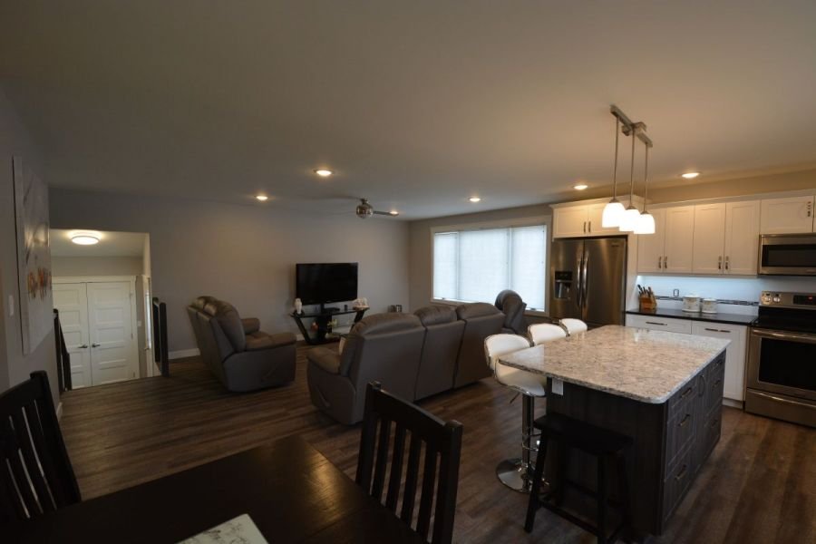 Open-concept living room and kitchen with beige walls, hardwood floors, a granite kitchen island, white cabinets, stainless steel appliances, a leather sectional sofa, and a dining table with four chairs.