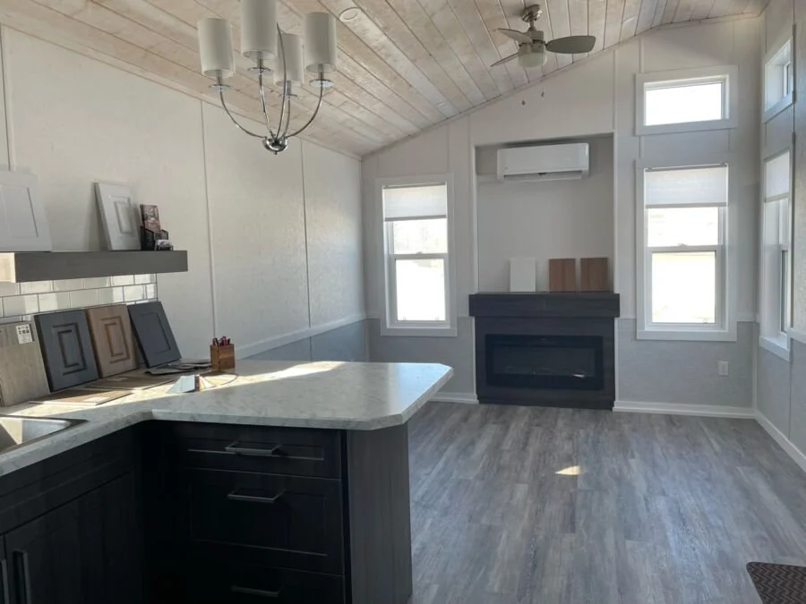 A modern, bright kitchen with white walls, a wooden ceiling, a chandelier, three windows, a fireplace, and a kitchen island with a marble countertop.