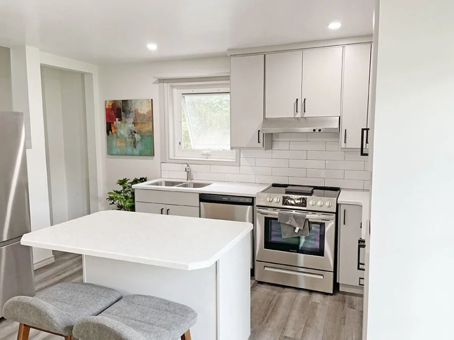 Modern kitchen with white cabinets, stainless steel appliances, a small countertop island, and a window over the sink, with a colorful abstract painting on the wall.