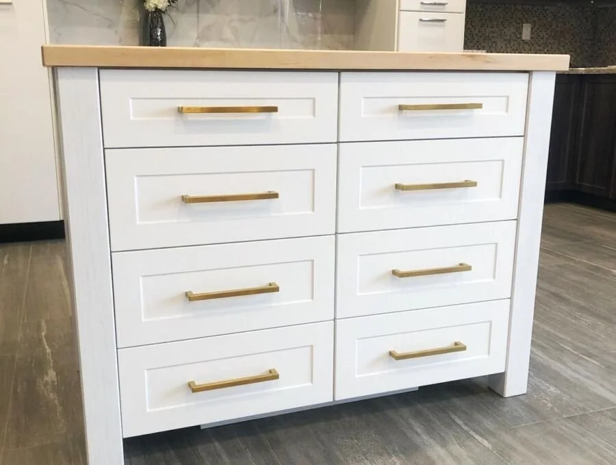 White kitchen island with six drawers, gold handles, and a light wood countertop in a modern kitchen.