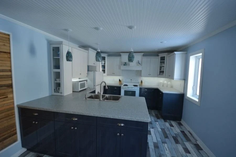 Modern kitchen with white upper cabinets, black lower cabinets, a gray speckled countertop, and a central island with a double sink. Wooden floors and a window on the right, with three pendant lights hanging above the island.