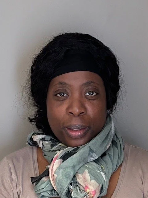 A woman with dark, curly hair wearing a black headband and a patterned scarf around her neck, photographed against a plain light gray background.