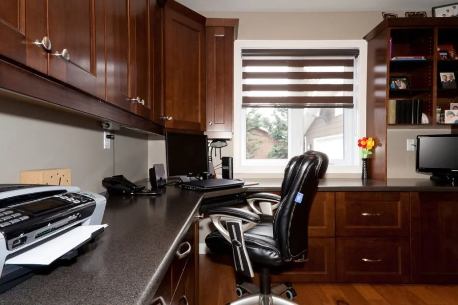 Home office with dark wood cabinets, a black desk with computers and office supplies, a window with striped blinds, and a vase with colorful flowers.