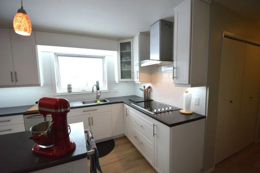 Modern kitchen with white cabinets, black countertops, a window above the sink, red stand mixer on the counter, and a paper towel holder, with light coming from a ceiling fixture.