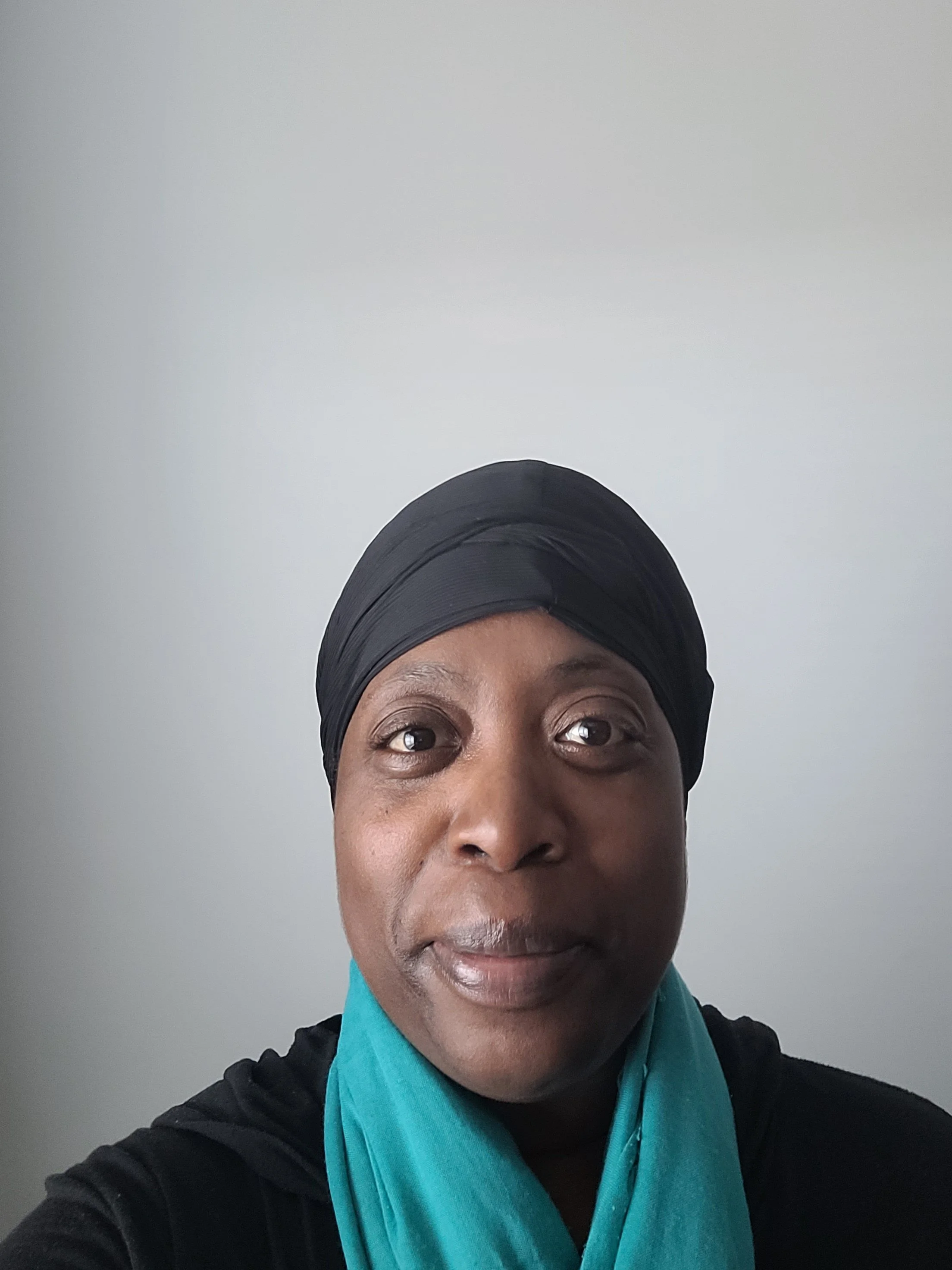 A woman with dark, curly hairwearing a black headband and a patterned scarf around her neck, photographed against a plain light gray background.