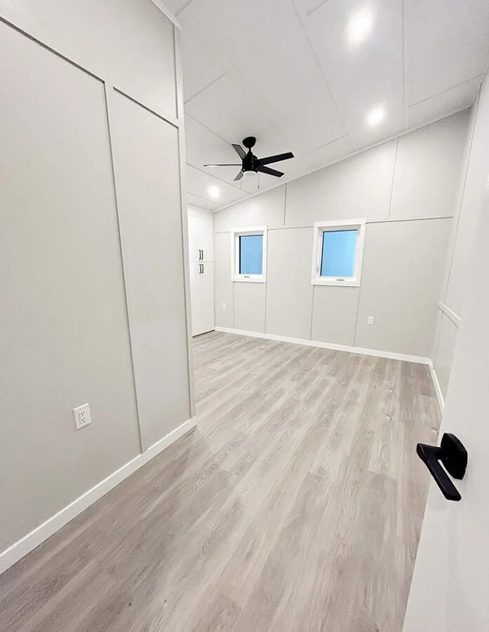 Empty room with light-colored walls and wood laminate flooring, two small windows, a ceiling fan, and a door with a black handle.