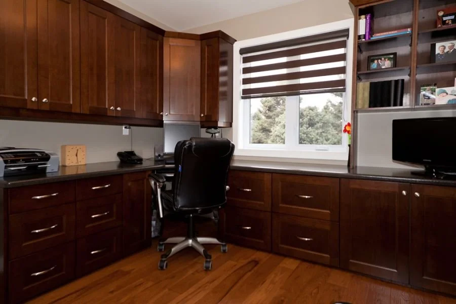 Home office with dark wood cabinets, a black leather office chair, a window with striped blinds, a computer monitor, a small decorative clock, and shelves with books and photos.