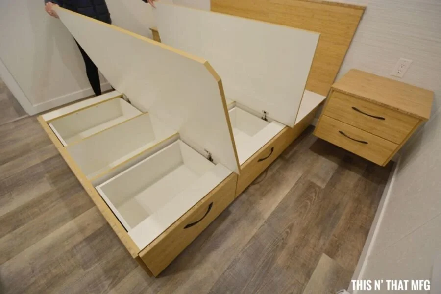 A person opening a partially assembled wooden bed with a built-in storage compartment at the foot, next to a matching wooden nightstand with two drawers, on a wooden floor in a bedroom.