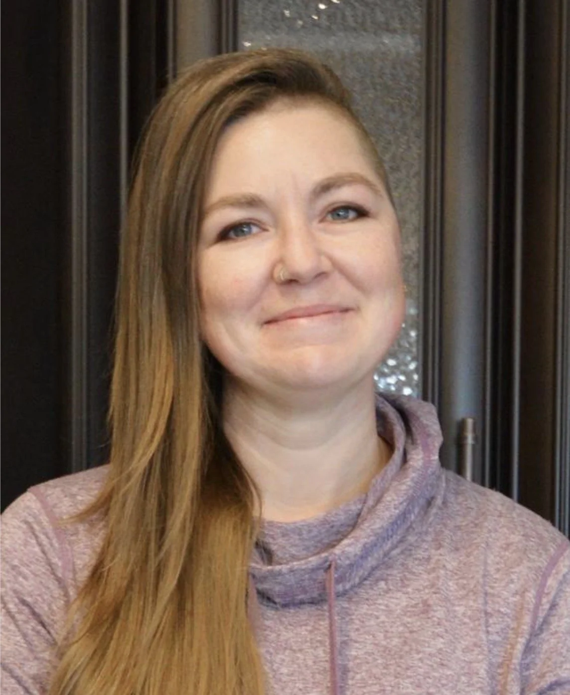 A woman with long brown hair and blue eyes smiling, wearing a gray athletic hoodie, standing in front of a dark wood-paneled background.