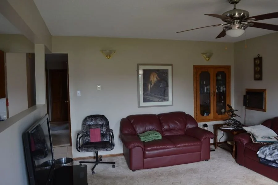 Living room with beige walls, a ceiling fan, two wall-mounted lights, a framed picture, maroon leather couch, a black office chair, glass cabinet, and a side table with various items.