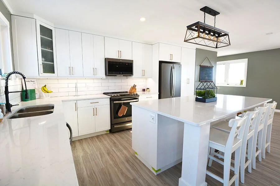 Modern white kitchen with an island, stainless steel appliances, and a light wood floor. There are four white chairs at the island and a hanging light fixture above.