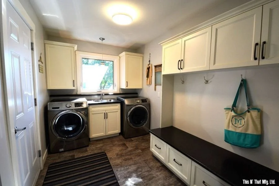 Laundry room with front-loading washer and dryer, white cabinets, window, black countertop, hooks on wall, and striped rug.
