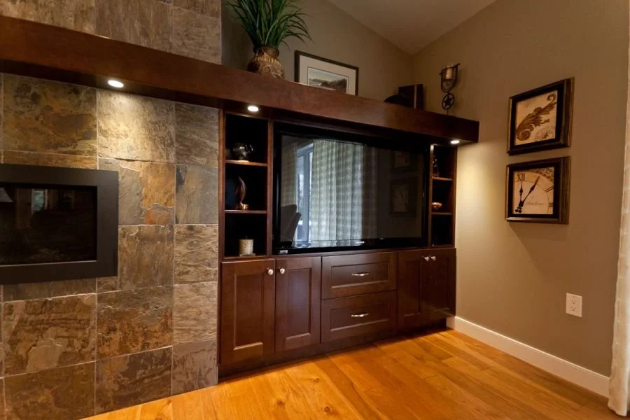 Living room with a flat-screen TV in a dark wood entertainment center, decorative shelves, framed artwork, a fireplace, and a potted plant on top. Hardwood floors and beige walls.