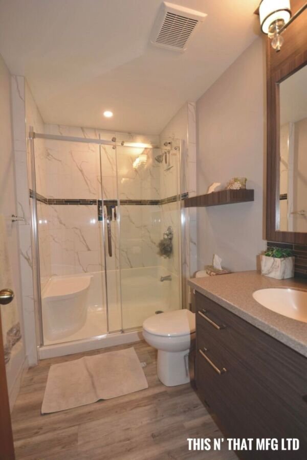 A bathroom with a glass-enclosed shower, a toilet, and a vanity with a sink. There is a mirror above the vanity, a tissue box, and a small shelf with decorative items. The floor is wooden and a white bath mat is in front of the shower.