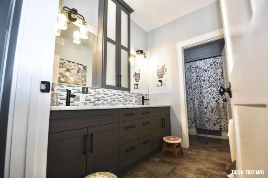 Bathroom with dark gray vanity, white and gray mosaic backsplash, framed mirror, wall sconces, a small stool with a patterned seat, and a doorway with floral curtains.
