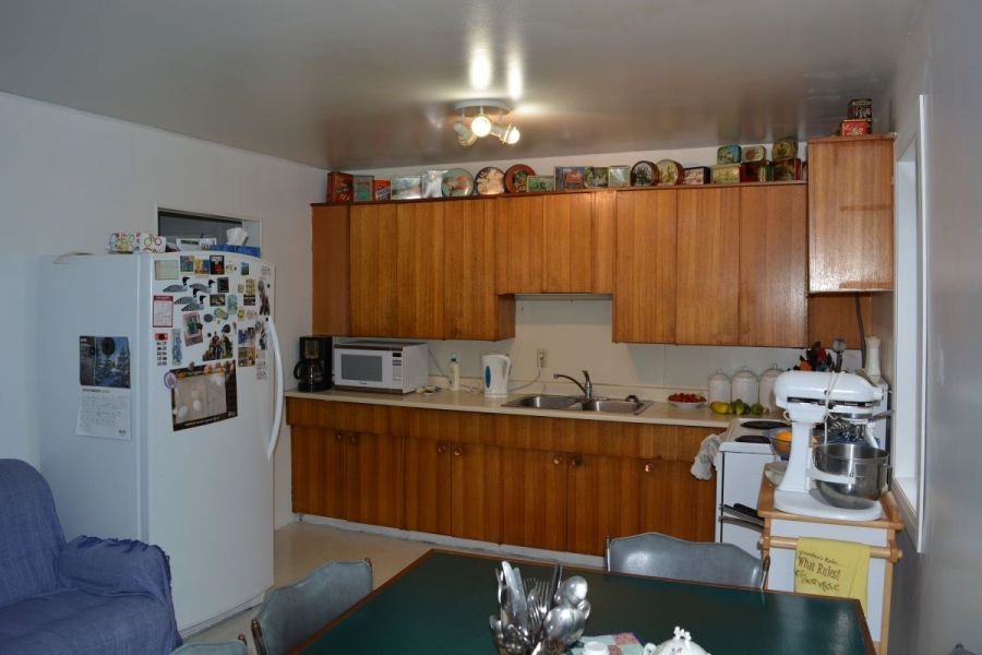 Kitchen with white refrigerator decorated with magnets, wooden cabinets, microwave, coffee maker, standing mixer, and some fruits on the counter. There is a dining table with chairs and a blue couch partially visible.