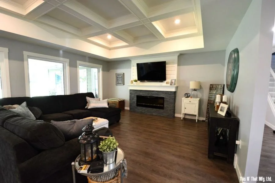 Living room with a large black sectional sofa, a gray stone fireplace with a mounted flat-screen TV above, and three windows letting in natural light. Decor includes a small white side table with a lamp, a black console table with framed photos, and wall decor. The ceiling features a tray design with recessed lighting. The floor is dark wood.