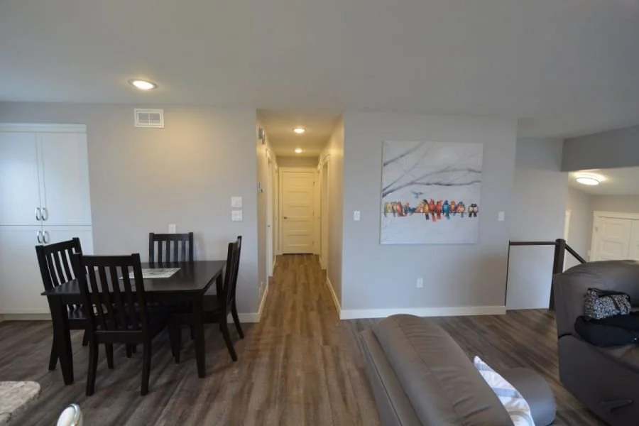Living room with a dining table and chairs, a gray couch, a colorful abstract painting on the wall, hardwood floors, and a hallway leading to a door.
