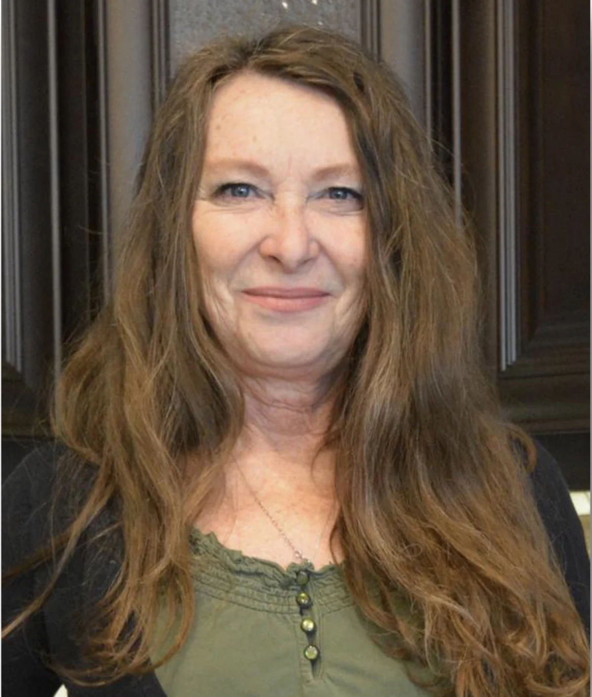 A woman with long, wavy brown hair, blue eyes, and a slight smile, wearing a green top and a black blazer, standing in front of dark wood cabinetry.