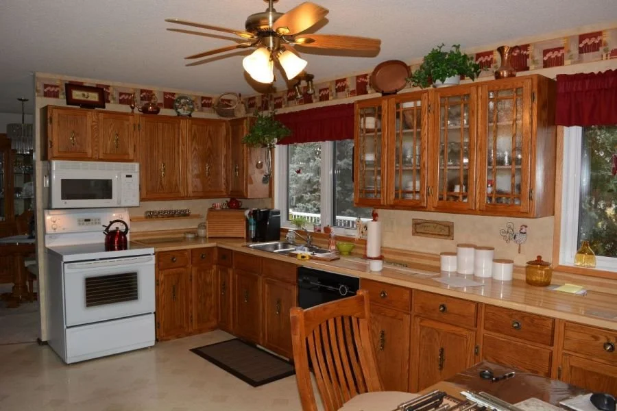 A traditional kitchen with wooden cabinets, a white stove, microwave, and black dishwasher, with a ceiling fan, large windows with red valances, and various kitchen items and decor.