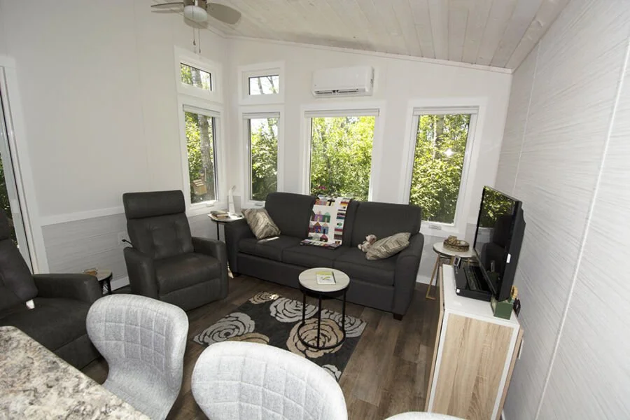 Living room with black leather recliner, gray sofa, round coffee table, TV on stand, multiple windows with views of trees, patterned rug, and white paneled walls.