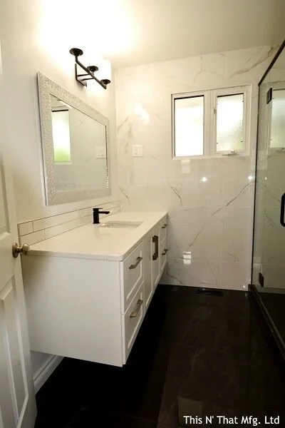 Bathroom with a white vanity, mirror, and black faucet, marble walls, a window, and a glass shower door.