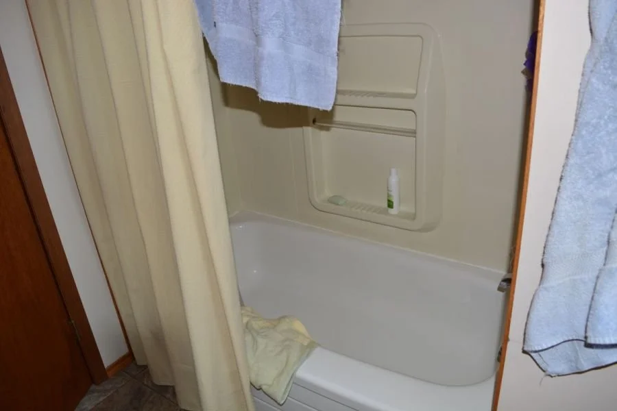 A small shower with a white bathtub, held back by beige shower curtains, with a blue towel hanging outside, a bottle of shower gel or shampoo on the built-in shelf, and a towel on the floor of the shower.