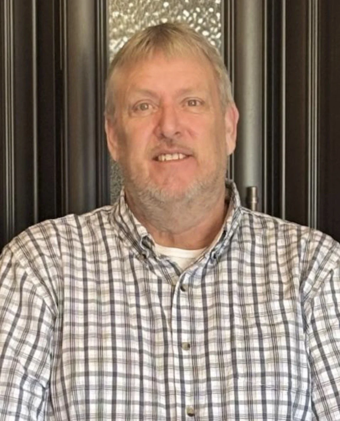 A middle-aged man with blond hair, beard, and mustache, smiling, wearing a plaid shirt, seated in front of a dark decorative wall.