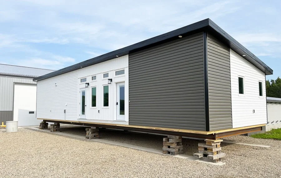 A small, modern, prefabricated house under construction, elevated on wooden supports, with white and dark gray siding.