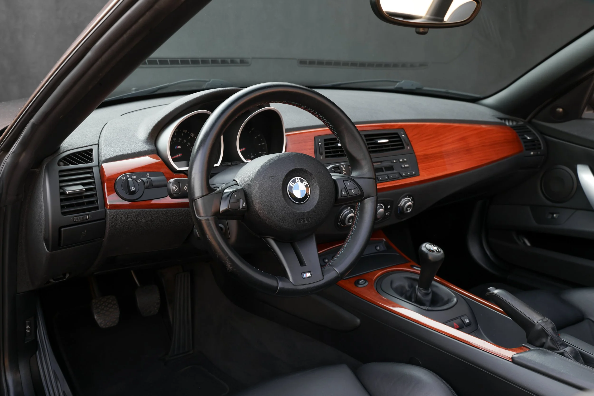 Interior of a BMW car showing the steering wheel, dashboard with tachometer and speedometer, and center console with controls, featuring wood trim accents.