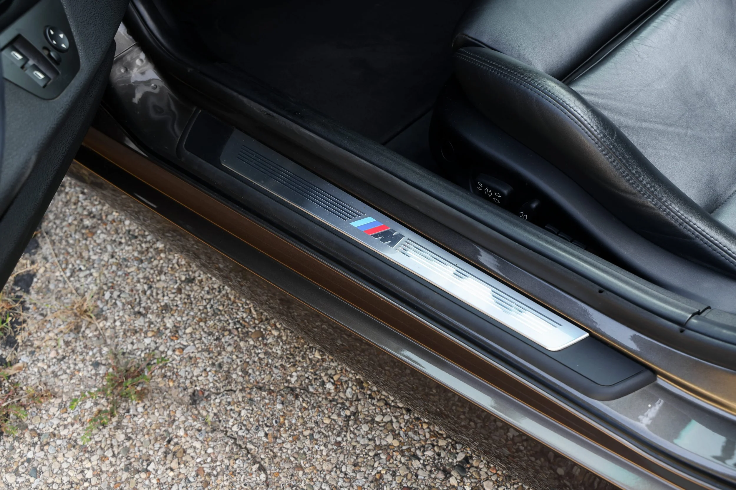 Close-up of a car door's sill with a metallic plate featuring the BMW M logo, black leather interior, and partially visible window controls against a rough gravel ground.