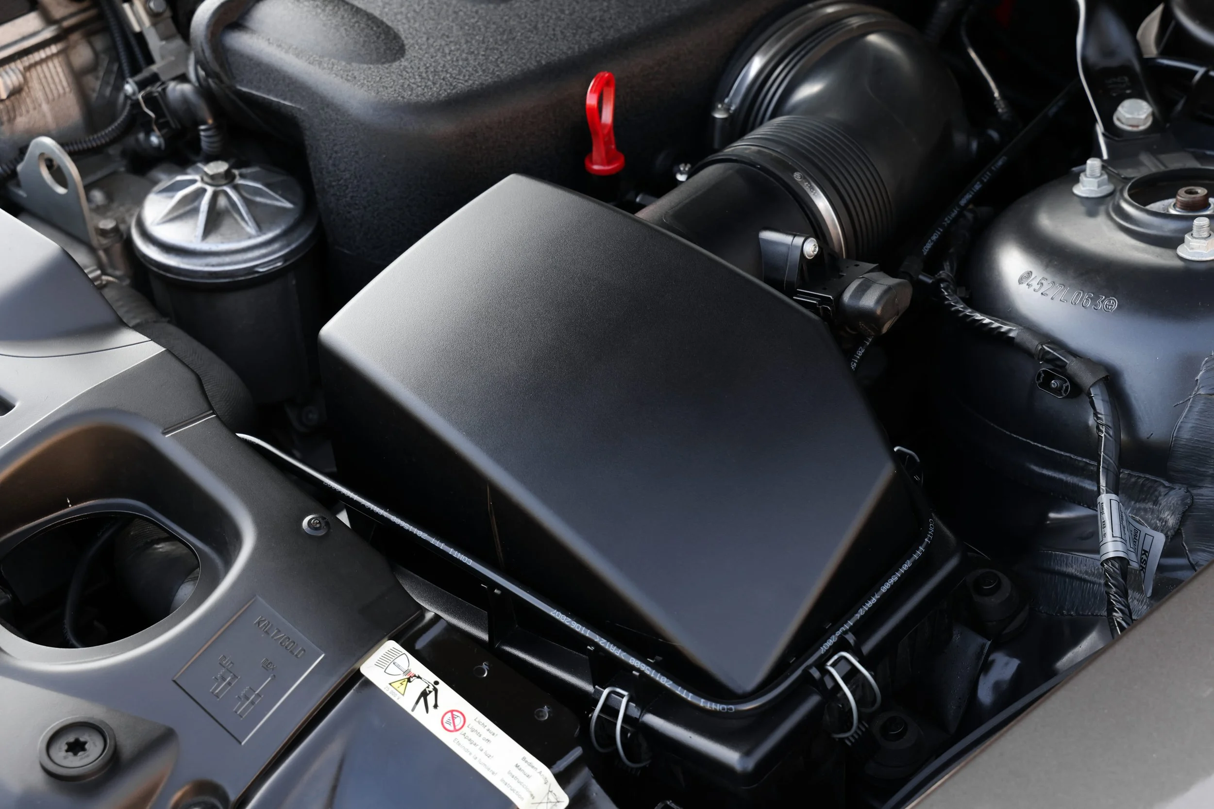 Close-up of a car engine showing various components including a black plastic cover, metallic parts, tubes, and warning labels.