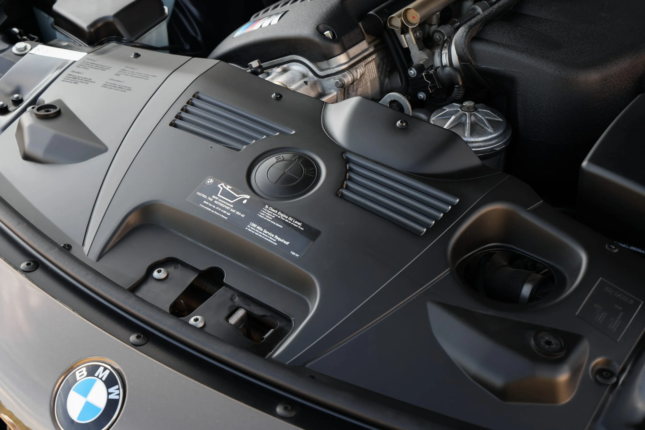 Close-up of a car engine with BMW and BMW Motorsport logos visible, showing various engine components and black cover plates.