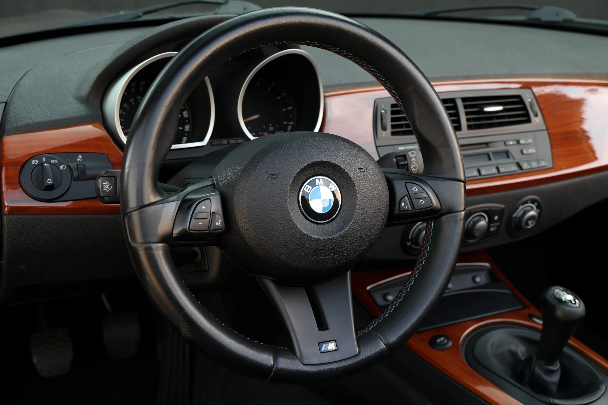 Interior of a BMW car showing the steering wheel with BMW emblem, dashboard with gauges, wood trim, and control buttons.