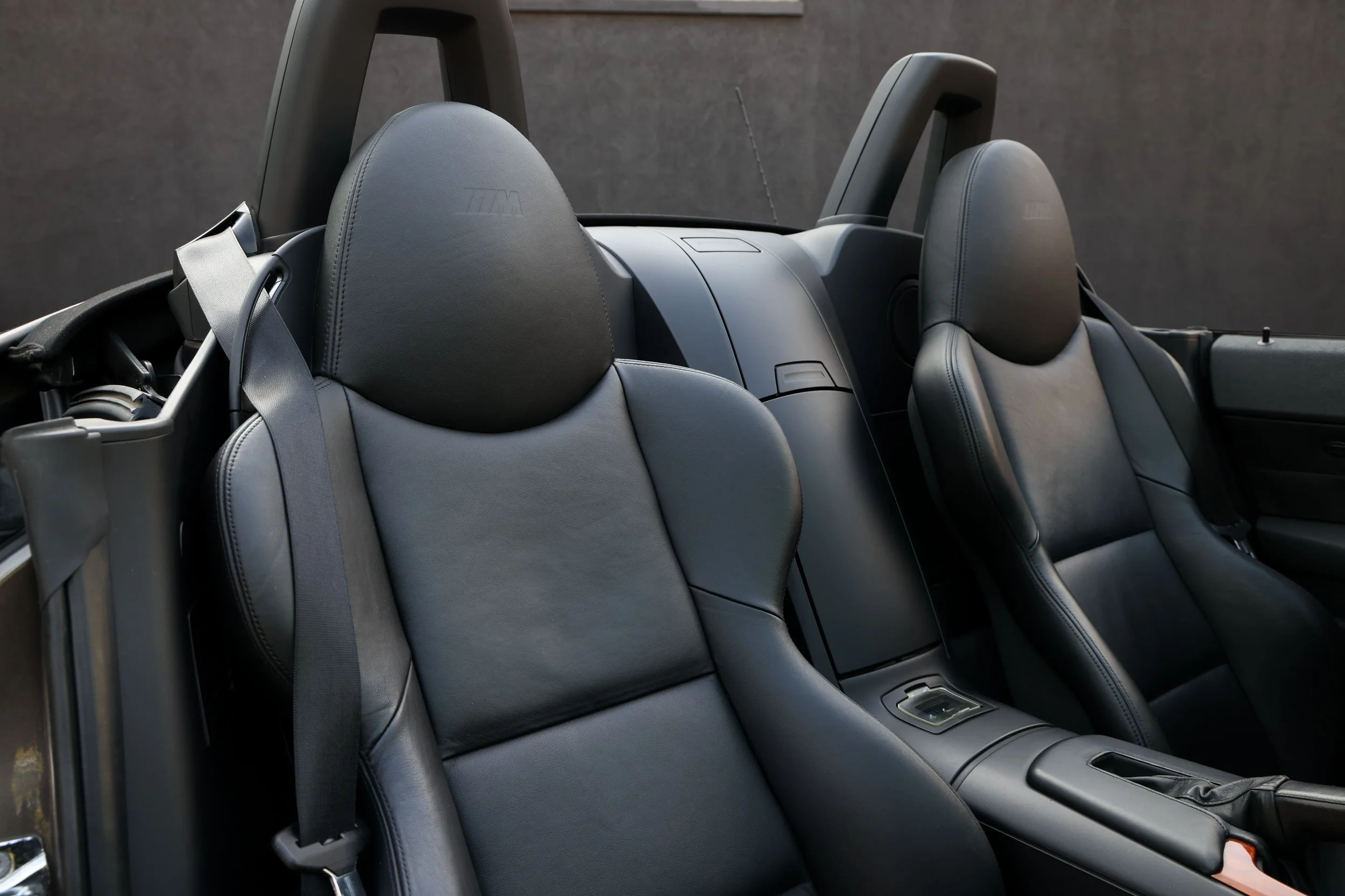The interior of a convertible car showing three black leather seats with the BMW M logo embossed on the headrests. The seats are equipped with seatbelts and the central console is visible in front of the back seat.