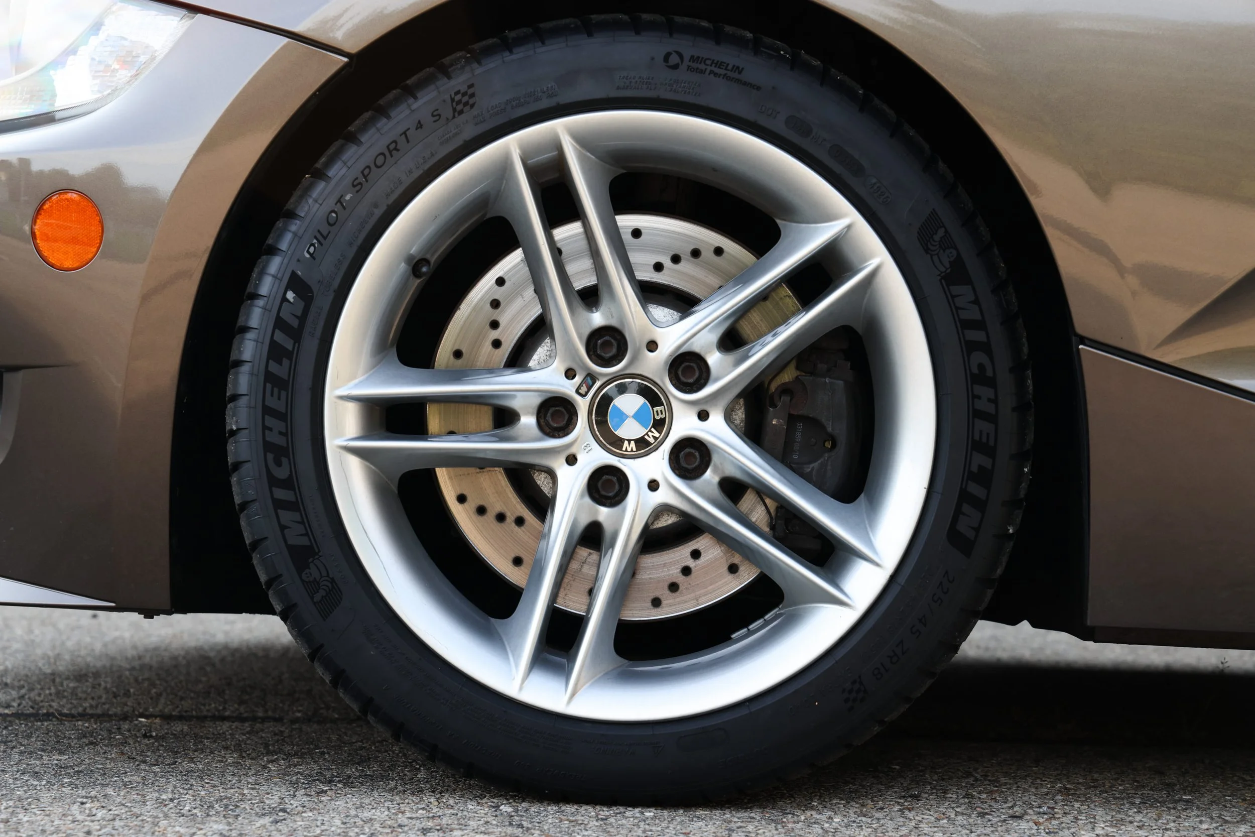 Close-up of a car's front wheel, featuring a silver alloy rim with a BMW logo at the center, a black tire with branding and specifications, and a brake disc behind the rim. Part of the car's beige body and a small orange reflector are visible.