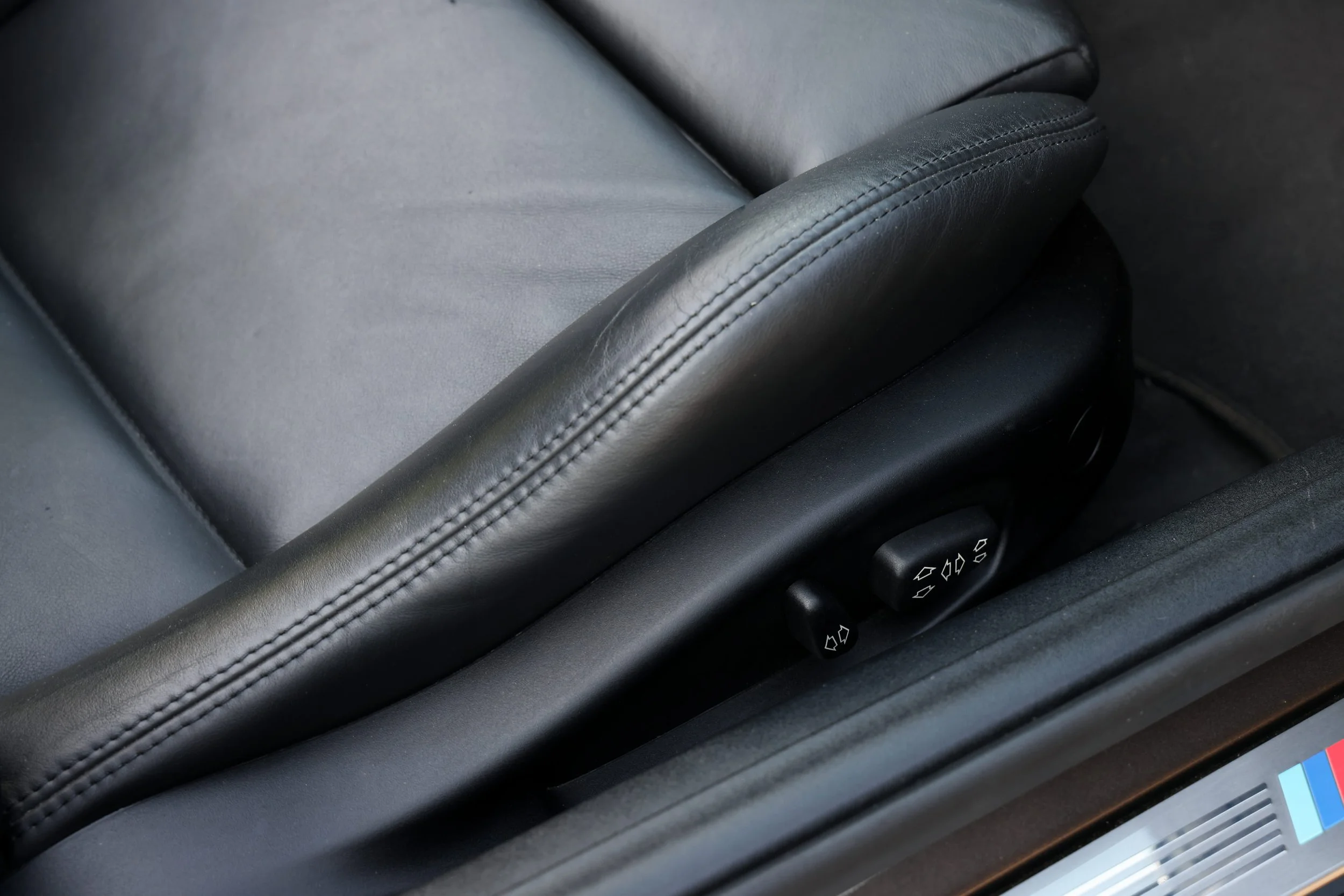 Close-up of a section of a car's black leather seat with stitching, a control panel with buttons, and part of a digital screen.