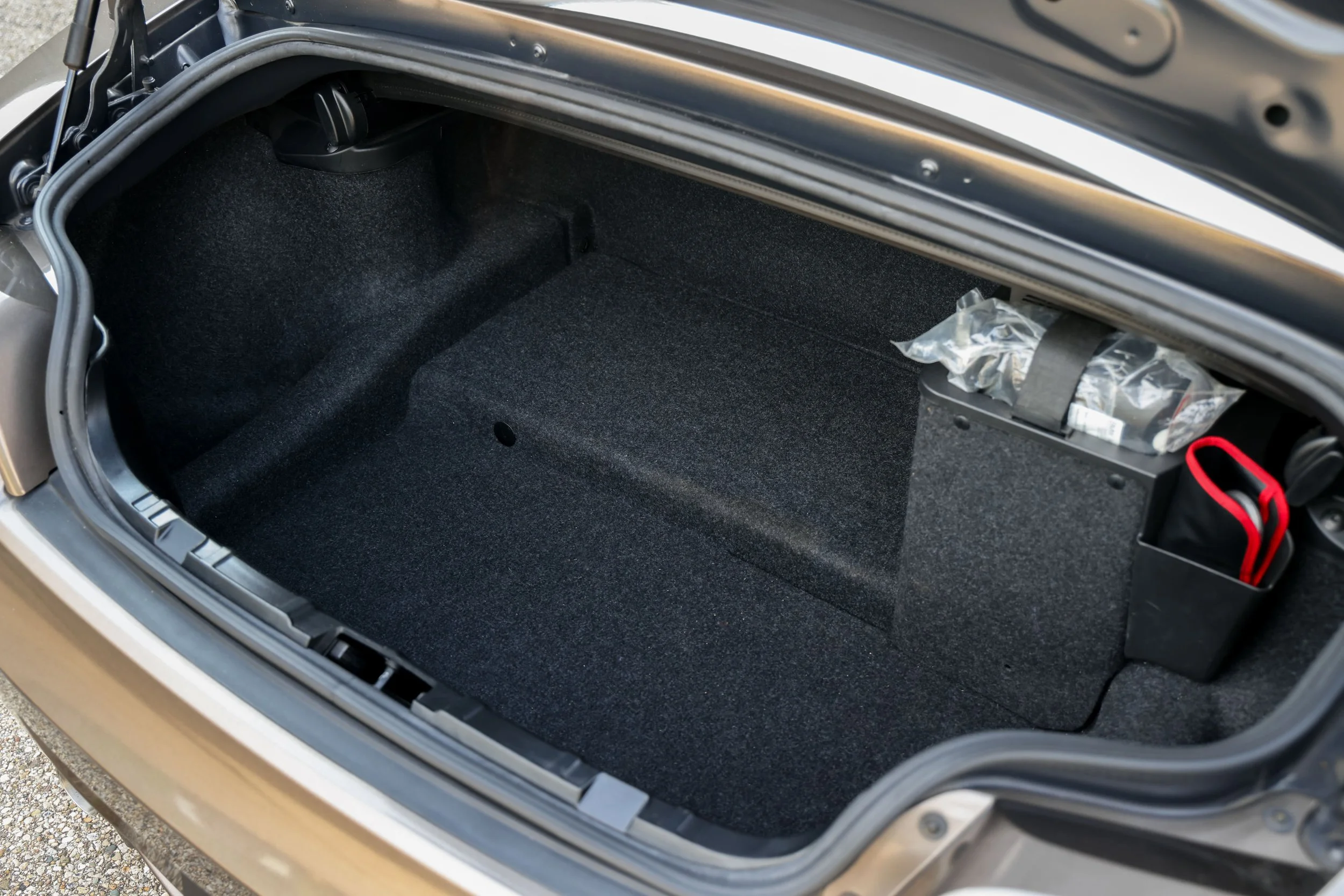 The image shows an open car trunk with a dark carpet lining inside. There is a small black object attached to the left side and a plastic bag in the right corner. The trunk is part of a silver-colored vehicle.