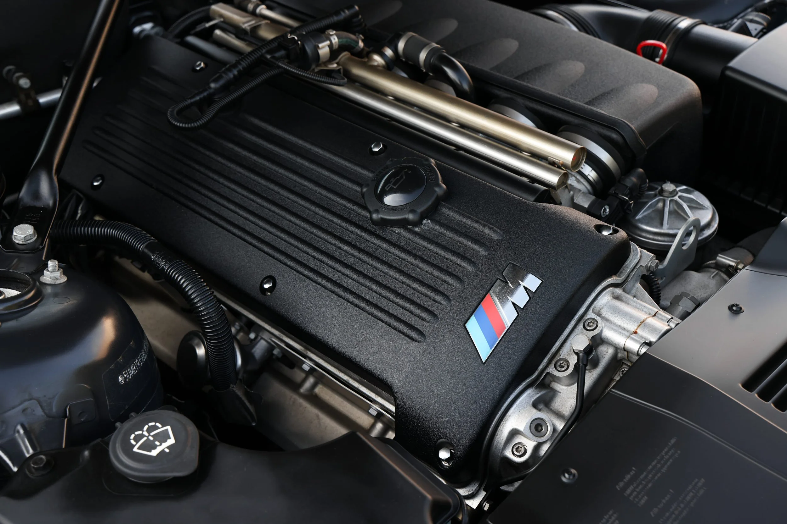 Close-up of a car engine with black cover, metallic pipes, and various components, including a BMW M logo.