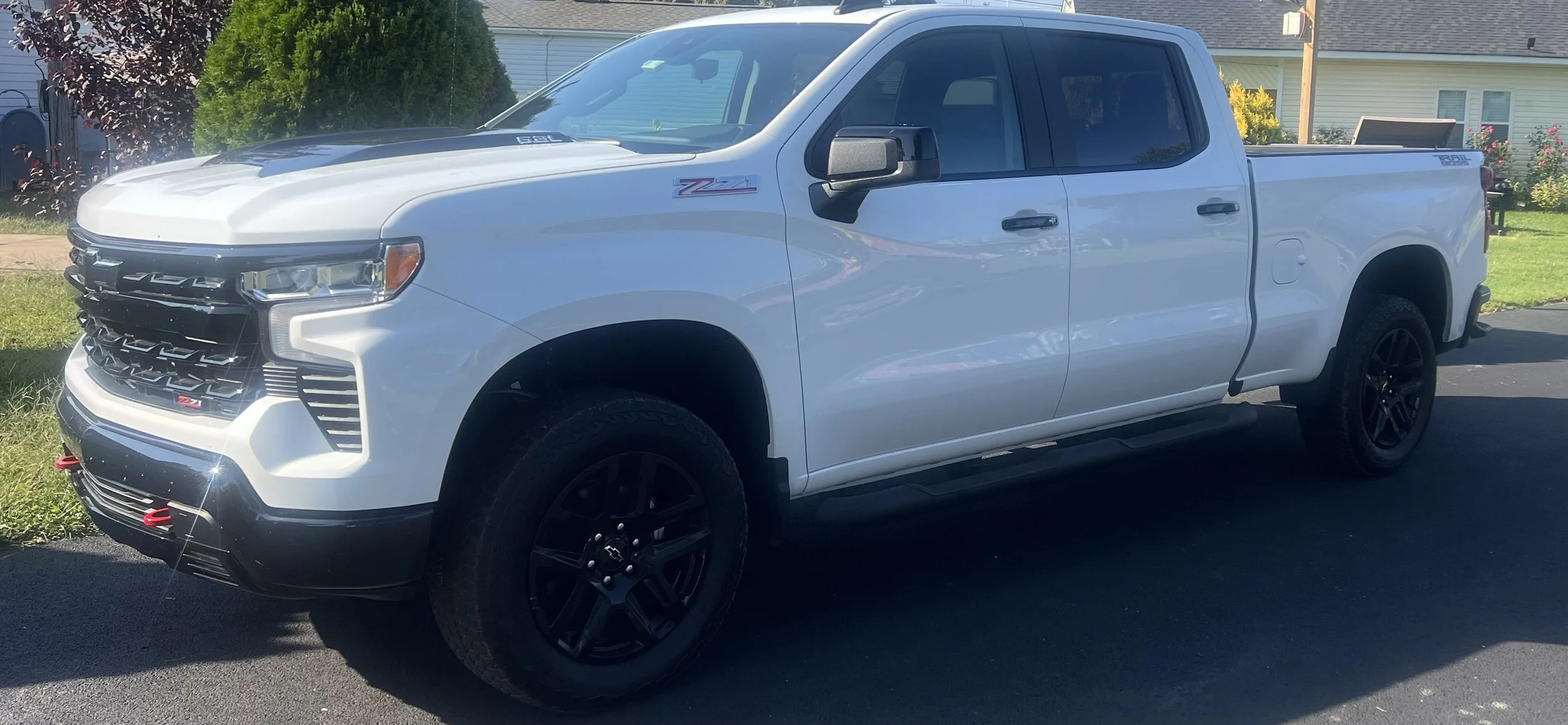 A white pickup truck with black wheels parked on a driveway in front of a house, with trees and bushes in the background.