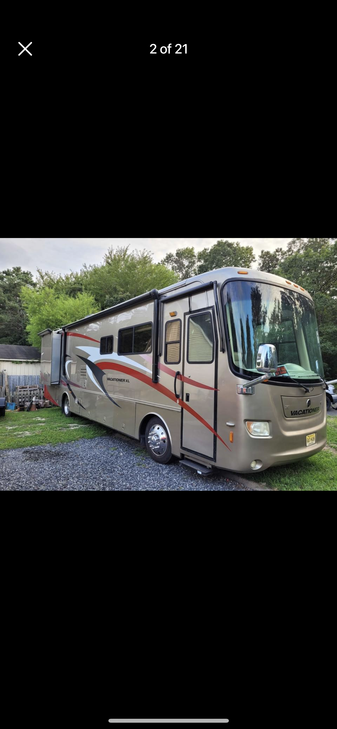 A beige vacationer XL motorhome parked on a gravel driveway surrounded by green trees and a wooden fence.