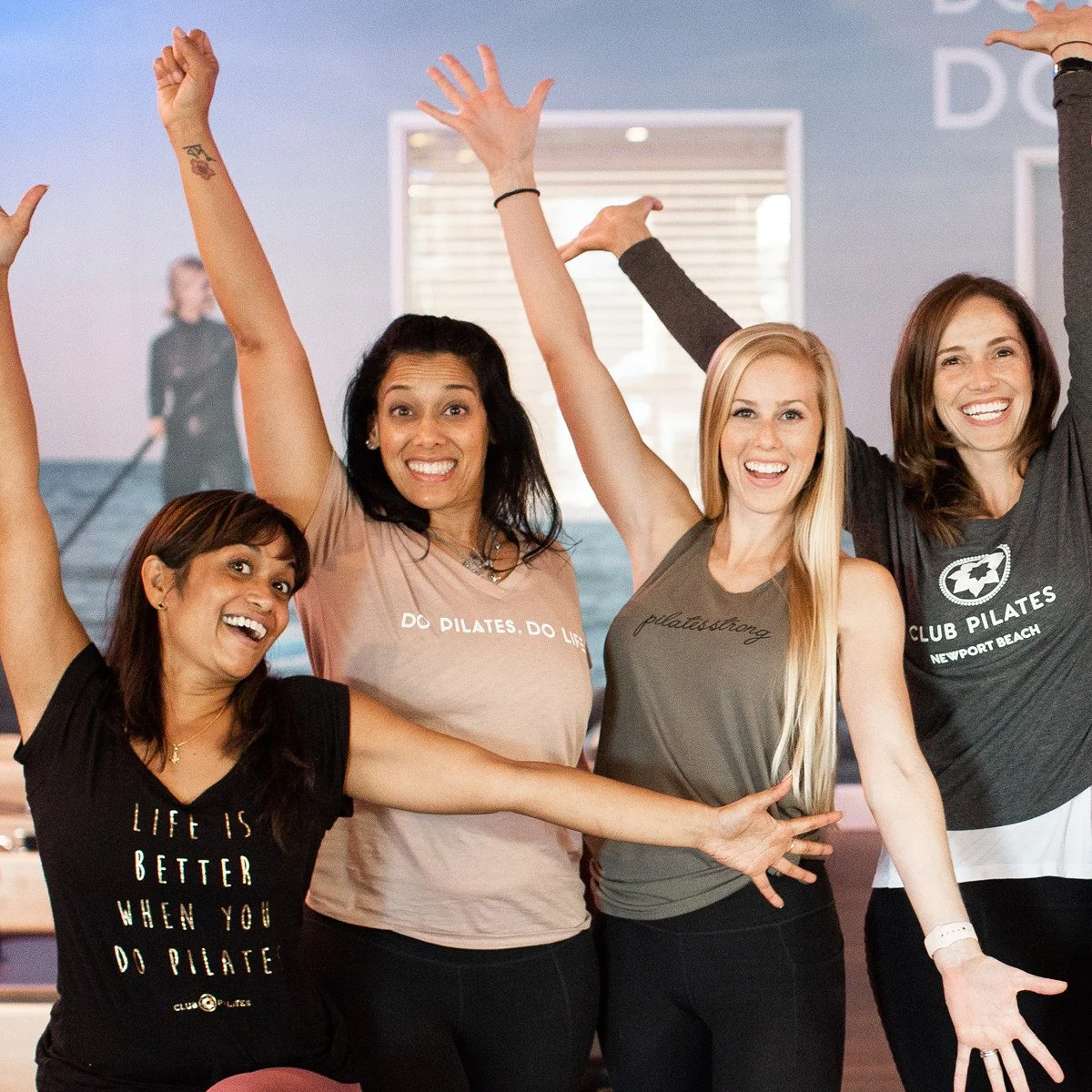 Four women smiling and posing with arms raised inside a fitness studio, with a woman in workout attire in the background on a paddleboard.