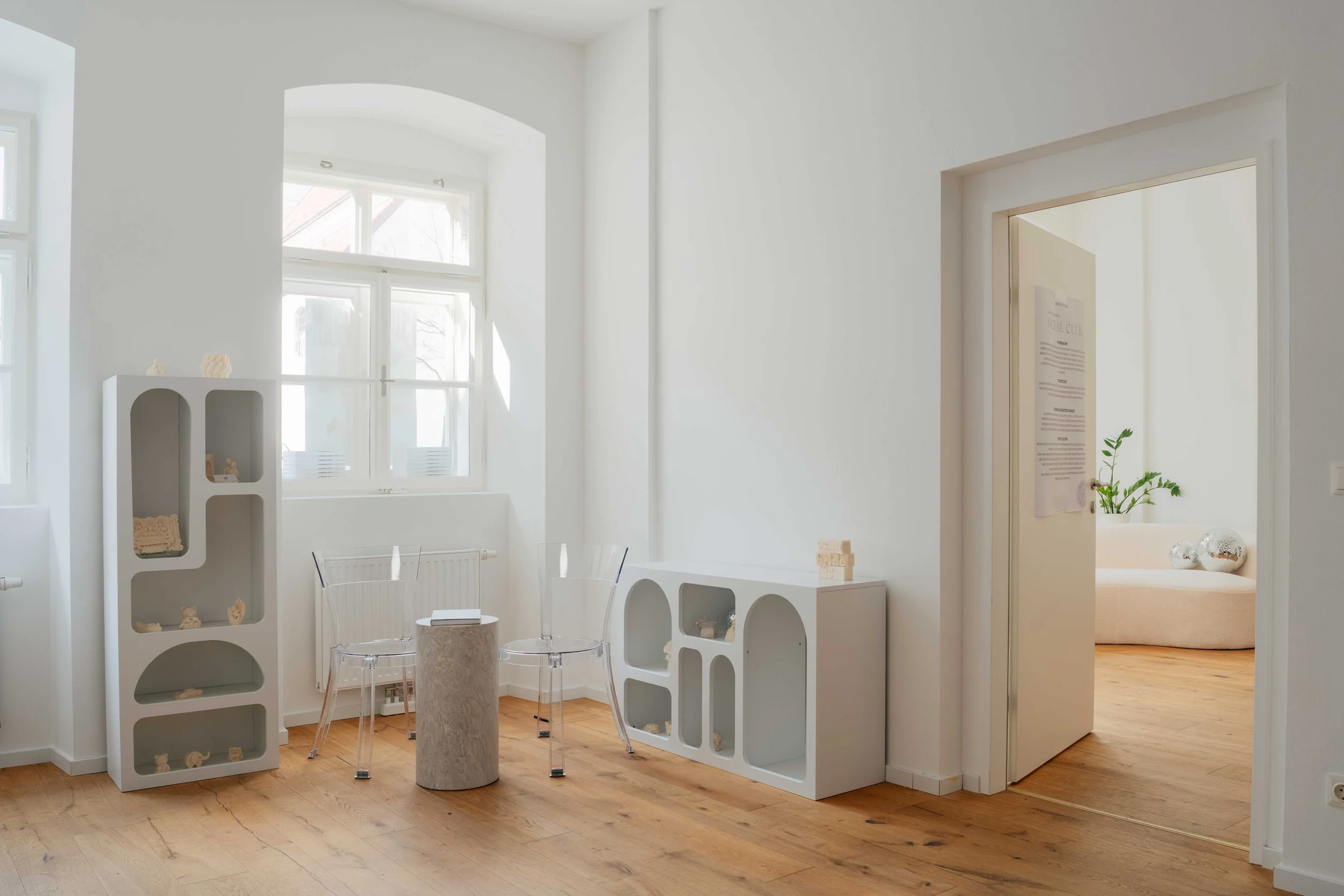 Ein heller Raum mit großen Fenstern, weißen Wänden und Holzboden. Es gibt moderne, Regale mit Dekorationsgegenständen und einen runden Beistelltisch mit transparenten Sesseln.