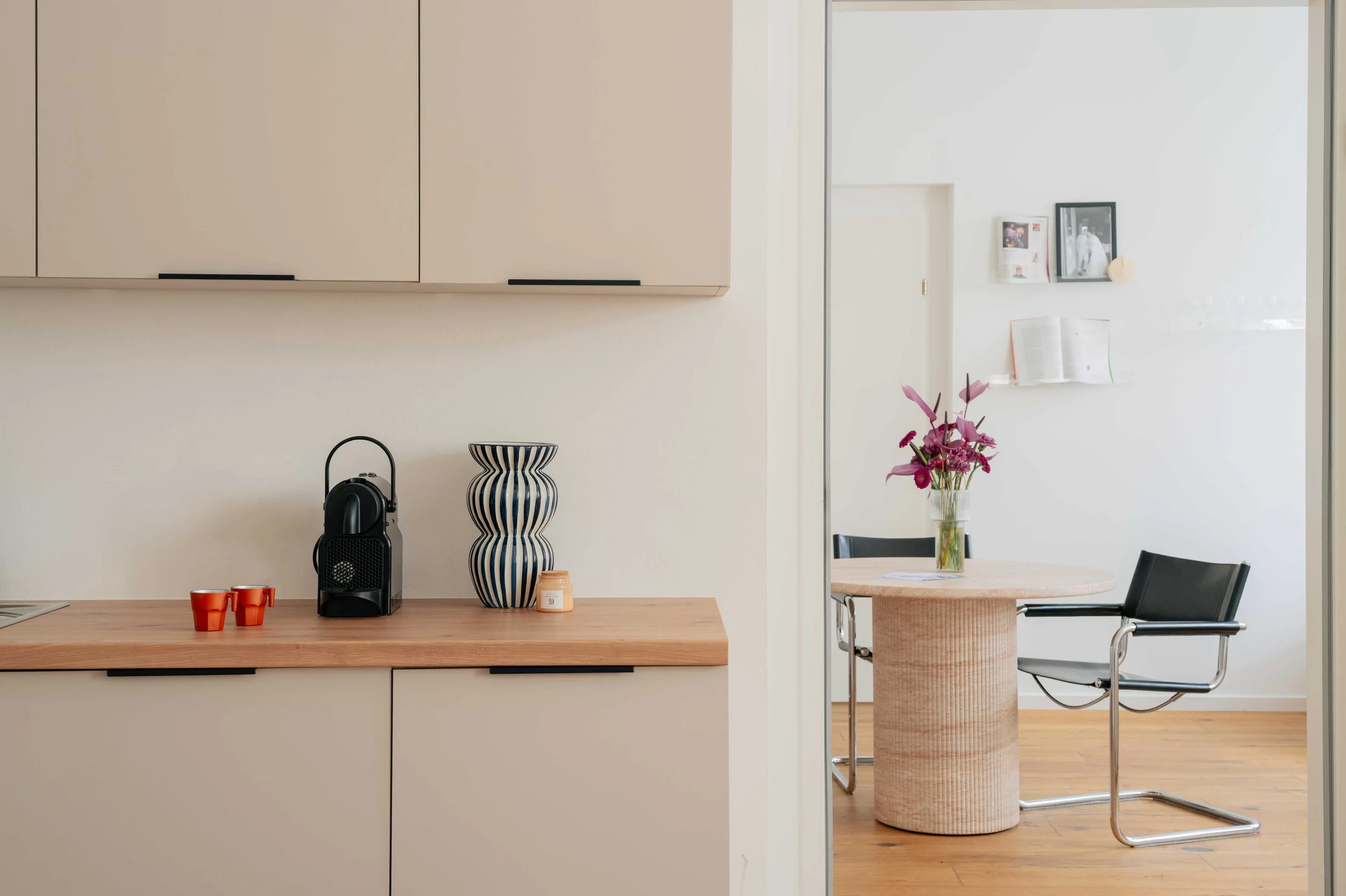 Innenraum einer modernen Küche und Esszimmer mit weißen Wänden, Holztisch und Stühlen, Vasen mit Blumen, Kühlschrank mit Fotos und Papieren, und einem hell erleuchteten Raum.