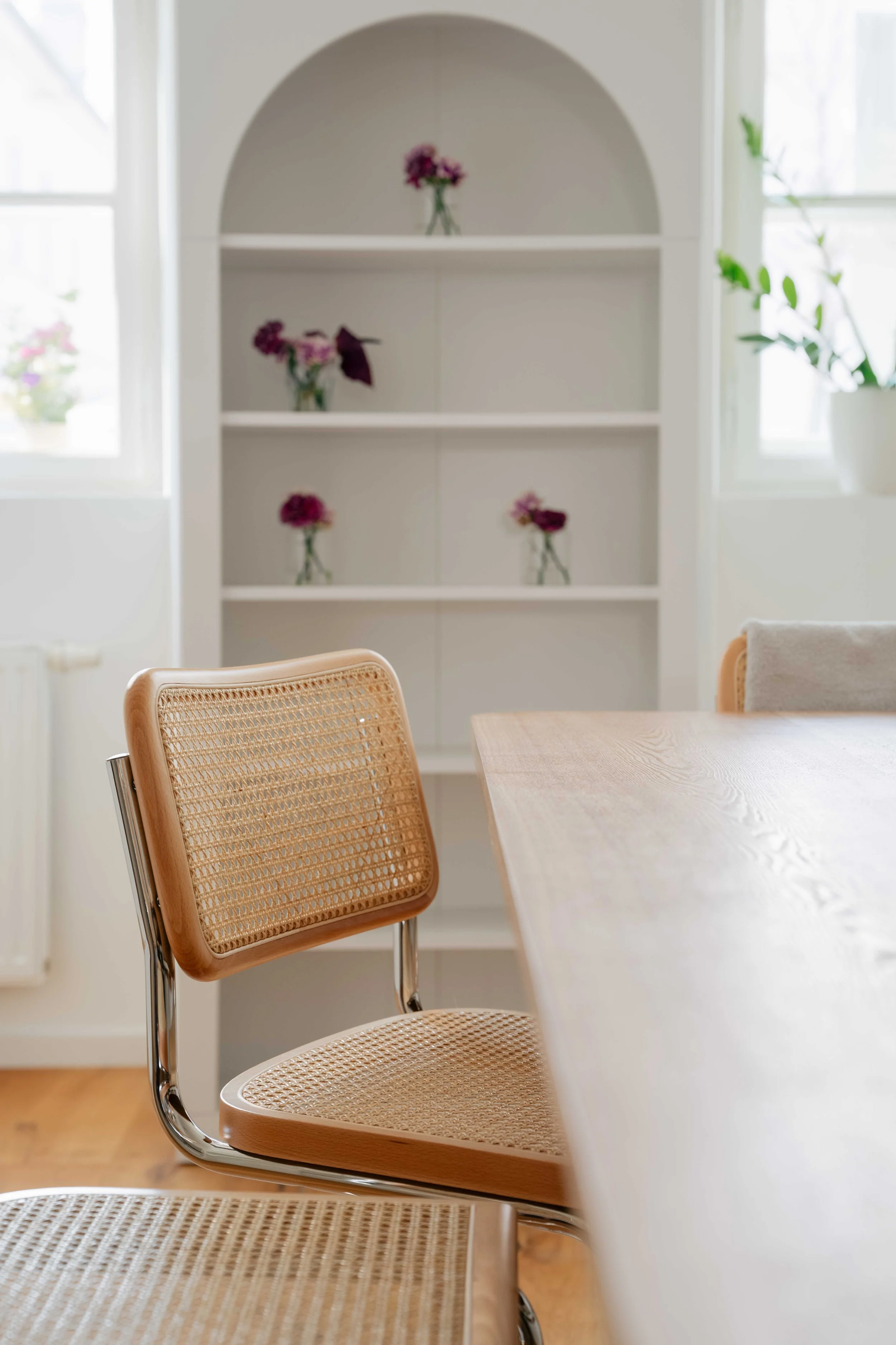 Ein heller Raum mit Fenster, einem weißen Bücherregal mit Blumenvasen und einem Holztisch mit hellen Rattan-Sesseln.