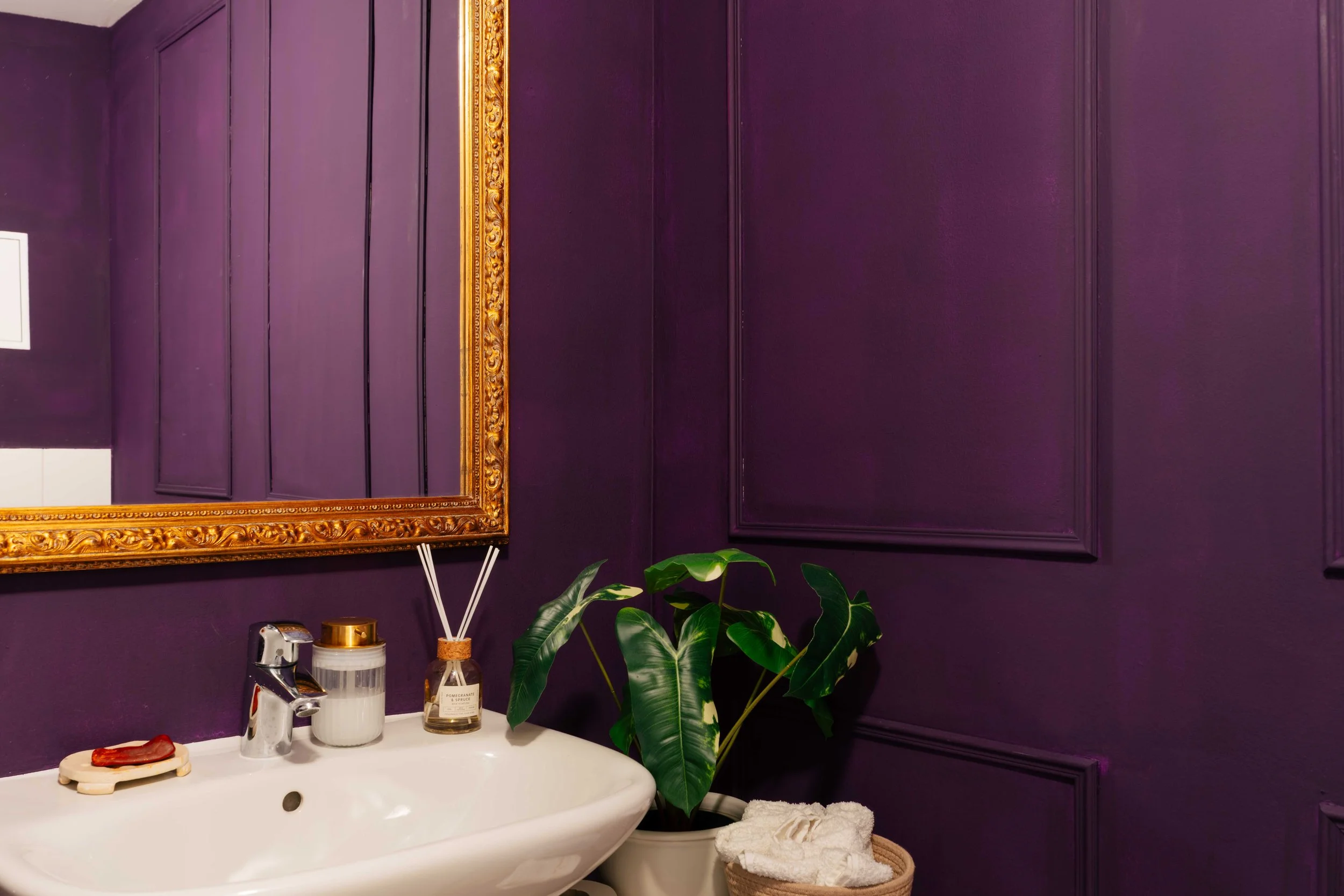Badezimmer mit dunkelvioletten Wänden, gold gerahmtem Spiegel, Pflanze, Waschbecken mit Seife, Kerzen und Handtüchern.