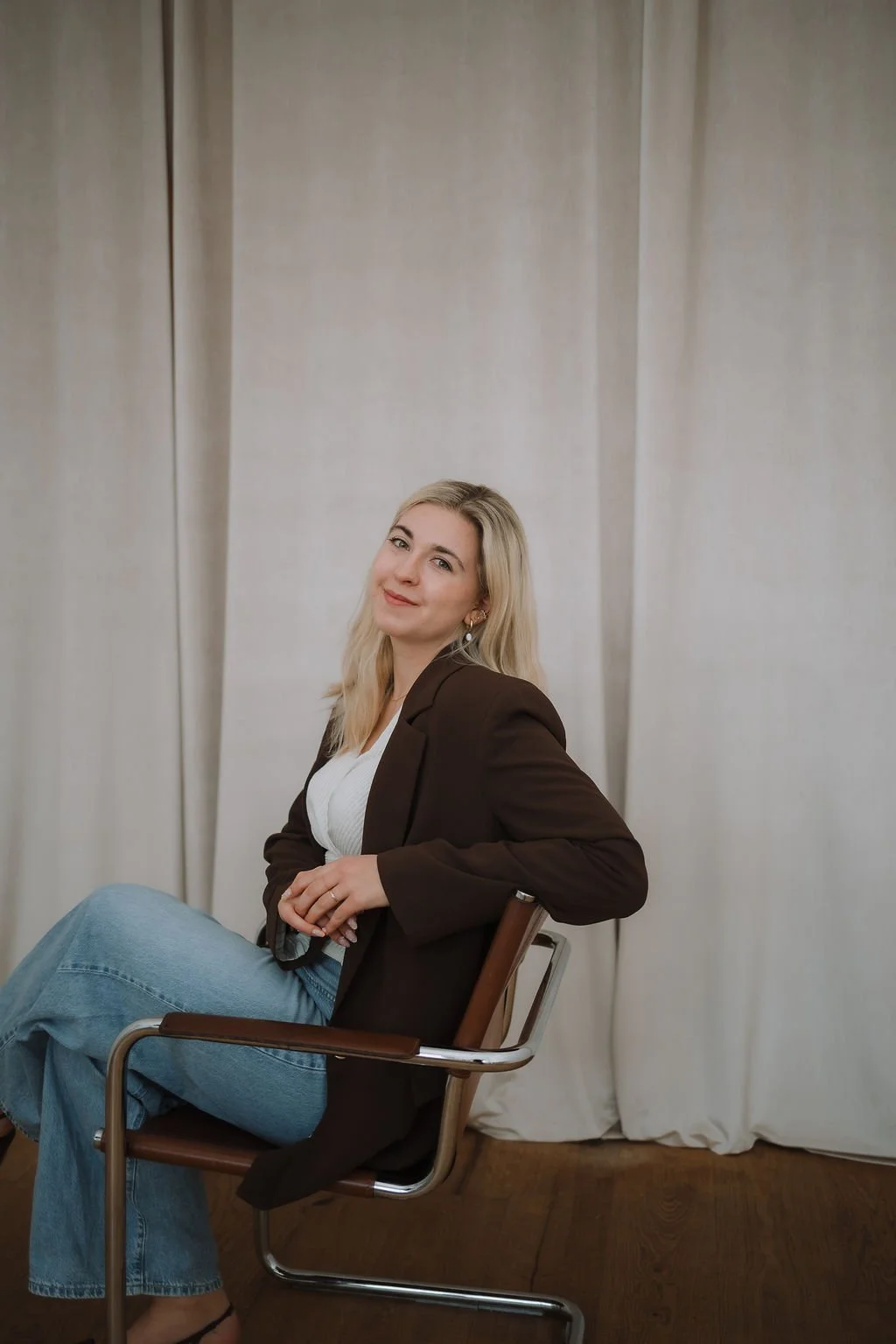 Eine lächelnde Frau mit blondem, lockigem Haar trägt eine weiß, gestreifte Wickel-Bluse, eine hellblaue Jeans und einen braunen Blazer und sitzt in einem Raum mit Holzboden auf einem braunen Sessel, vor einem hellen Stoffhintergrund.