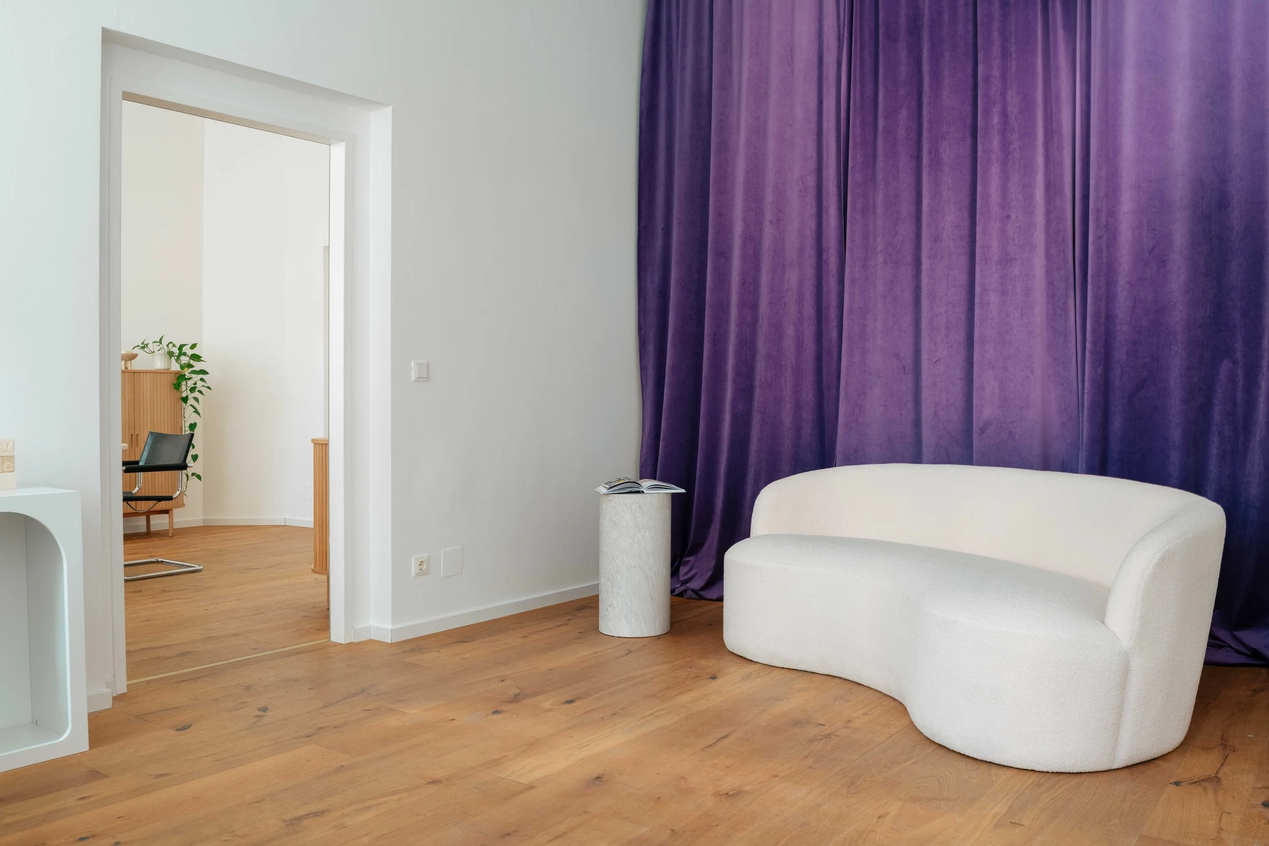Blick in ein Zimmer mit weißer Wand, violetter Vorhang, weißem Sessel und einem kleinen Beistelltisch mit einer offenen Zeitschrift darauf, daneben eine Tür zu einem weiteren Raum, in dem ein Schreibtisch und ein Hocker mit einem Pflanzentopf stehen.