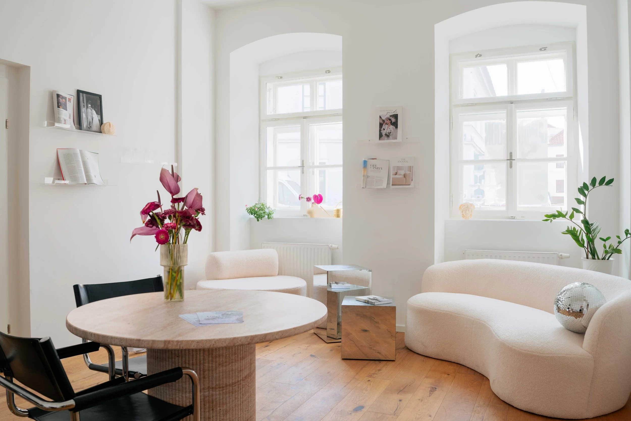 Heller, moderner Wohnraum mit großen Fenstern, weißen Wänden, einem runden Esstisch mit Blumen, einer weißen Couch, einem Sessel, und Dekorationsgegenständen auf den Fensterbrettern.